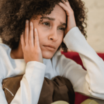 Woman sitting on couch holding pillow up to chest with hands on head and face looking anxious and worried