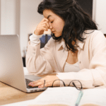 Woman with dark hair with her head in her hand, glasses on desk and computer open in front of her, showing suppressed anger in adults