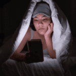 woman with duvet over her head in the dark on her phone unable to sleep and looking up treatment of insomnia in adults