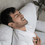 adult man sitting on the sofa in pain with his hands clutching his heart due to heart palpitations & anxiety in adults