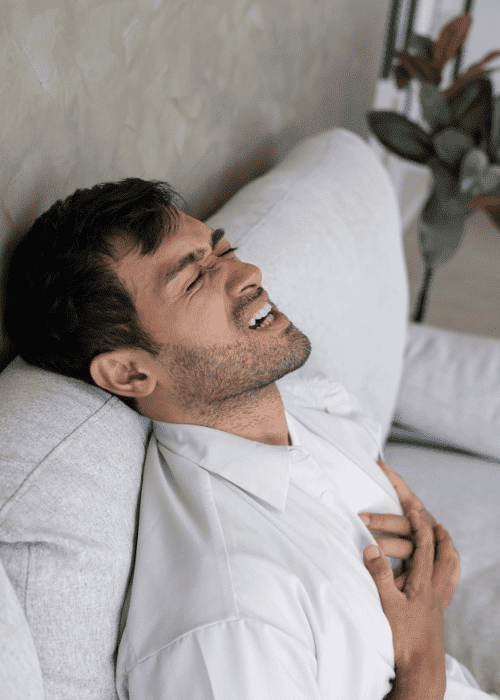 adult man sitting on the sofa in pain with his hands clutching his heart due to heart palpitations & anxiety in adults