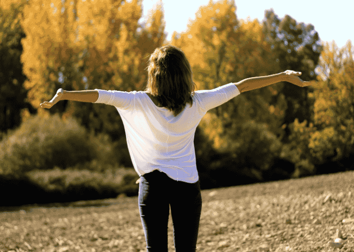 Woman standing in a park looking at trees with her arms up after getting treatment for nausea from stress or anxiety
