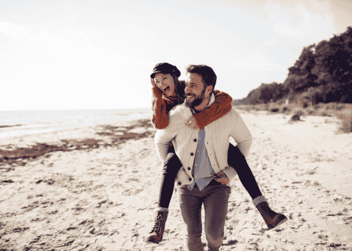 Happy couple with man giving woman a piggyback on the beach after receiving treatment for codependency in adults