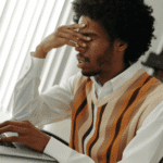man sitting at desk working on laptop with hand over eyes due to experiencing stress-induced forgetfulness in adults