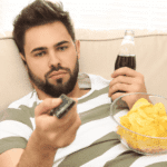 man on sofa drinking fizzy drink and balancing bown of chips on stomach whilst holding the remote avoiding responsibilities in adults