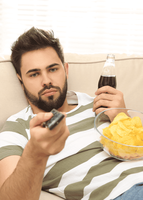 man on sofa drinking fizzy drink and balancing bown of chips on stomach whilst holding the remote avoiding responsibilities in adults
