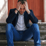 man sitting outside on step in front of door with head in hands due to compromised judgment under stress.