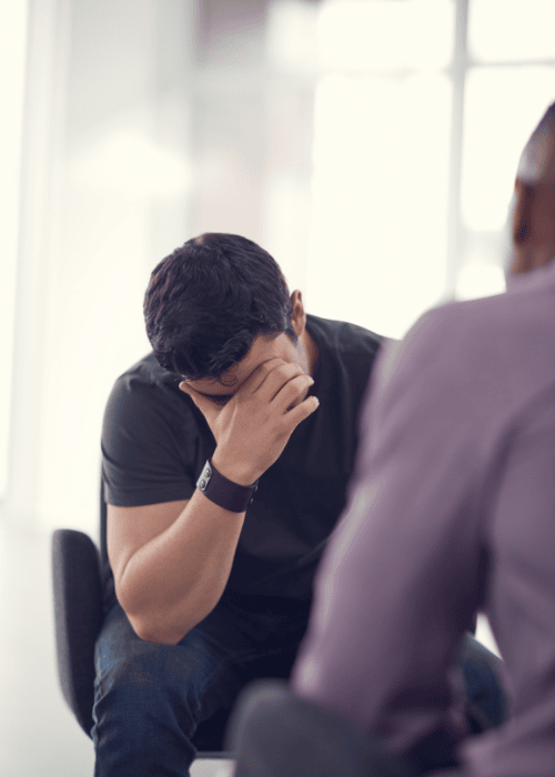 Man sitting across from a therapist with his head in his hands talking about overcompensation in adult relationships