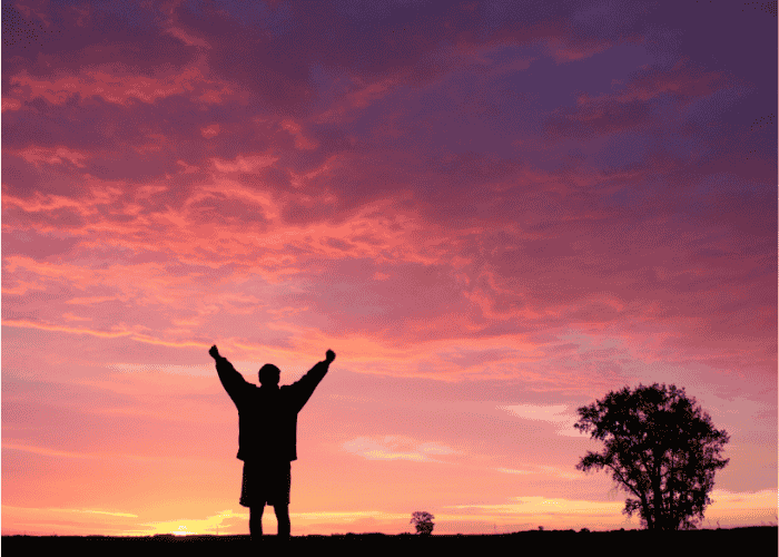 Man standing in front of a sunset holding his hands up in relief after receiving treatment for nervous fidgeting in adults