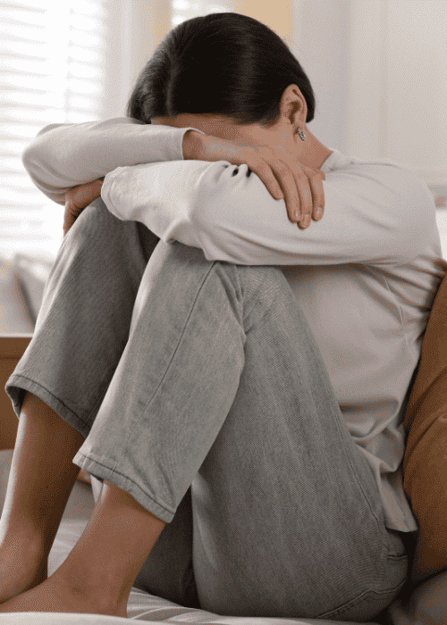 Women sitting on sofa with knees drawn to chest and face buried in arms worrying about Carelon Behavioral Health Insurance