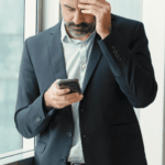 man wearing suit standing in office looking at phone rubbing forehead with his hand worrying about ComPsych mental health coverage