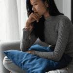 woman sitting cross-legged on sofa with cushion on lap and resting chin on hand worrying about First Health Network mental health insurance coverage