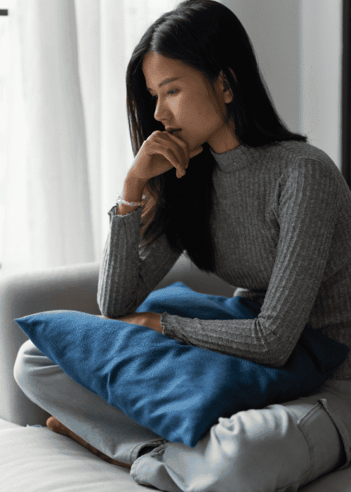 woman sitting cross-legged on sofa with cushion on lap and resting chin on hand worrying about First Health Network mental health insurance coverage