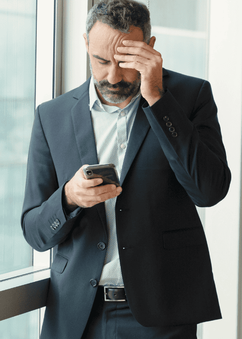 man wearing suit standing in office looking at phone rubbing forehead with his hand worrying about ComPsych mental health coverage