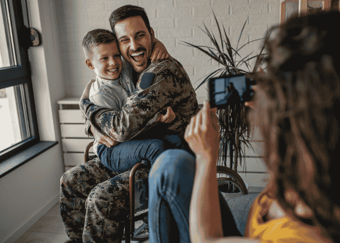 Male veteran in wheelchair with son sitting on his lap while mom takes a photo on her phone, smiling after seeking support with TriWest coverage for mental health treatment