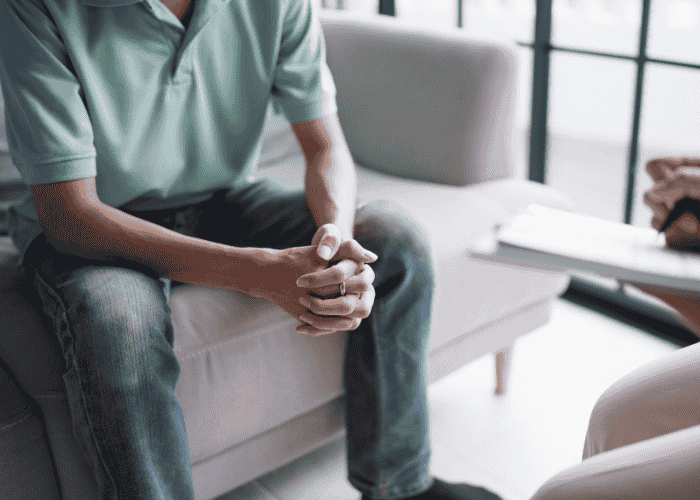 Man sitting on sofa with hands together and elbows resting on thighs opposite a therapist after seeking support using insurance to cover mental health treatment