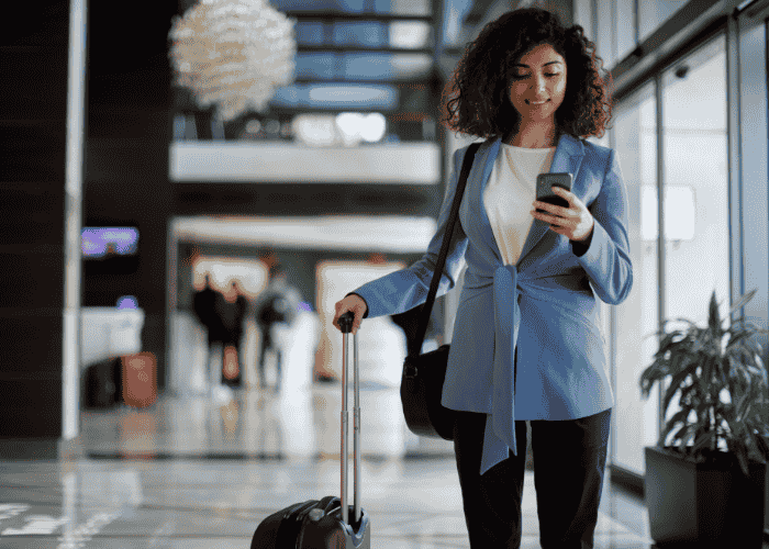 Woman with suitcase in airport terminal looking at phone and smiling after support with telehealth access for traveling professionals