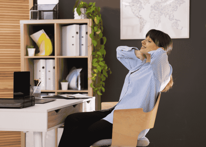 Woman sitting at desk leaning back with arms behind her head smiling after support with workplace reintegration after leave