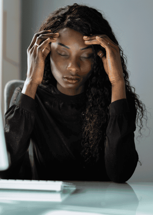 Woman sitting at desk with laptop with hands on head and eyes closed needing support with HPA axis dysfunction