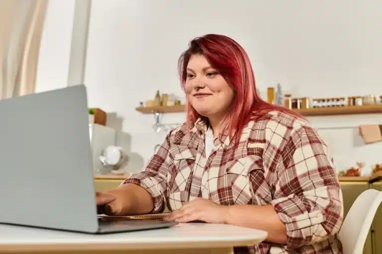 Woman with red hair wearing a flannel sitting at a desk looking at a computer during virtual telehealth outpatient mental health treatment