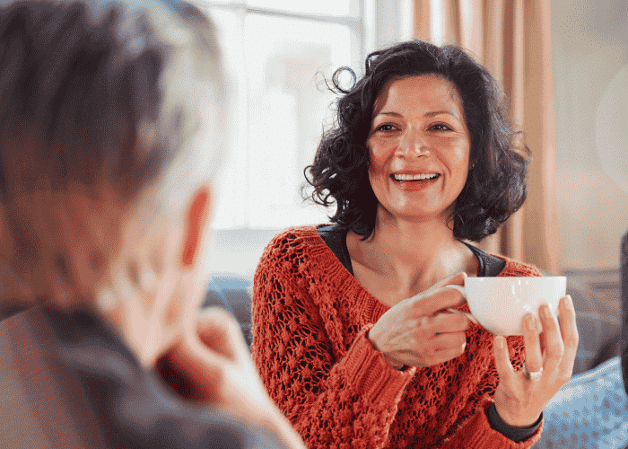 Woman sitting on couch with friends holding a cup of tea and smiling after support with perimenopause and mental health