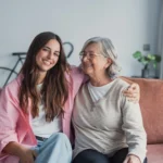 Adult daughter sitting on couch with arm around her mother smiling
