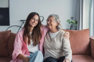 Adult daughter sitting on couch with arm around her mother smiling