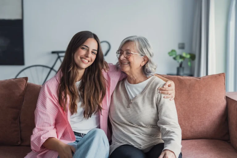 Adult daughter sitting on couch with arm around her mother smiling